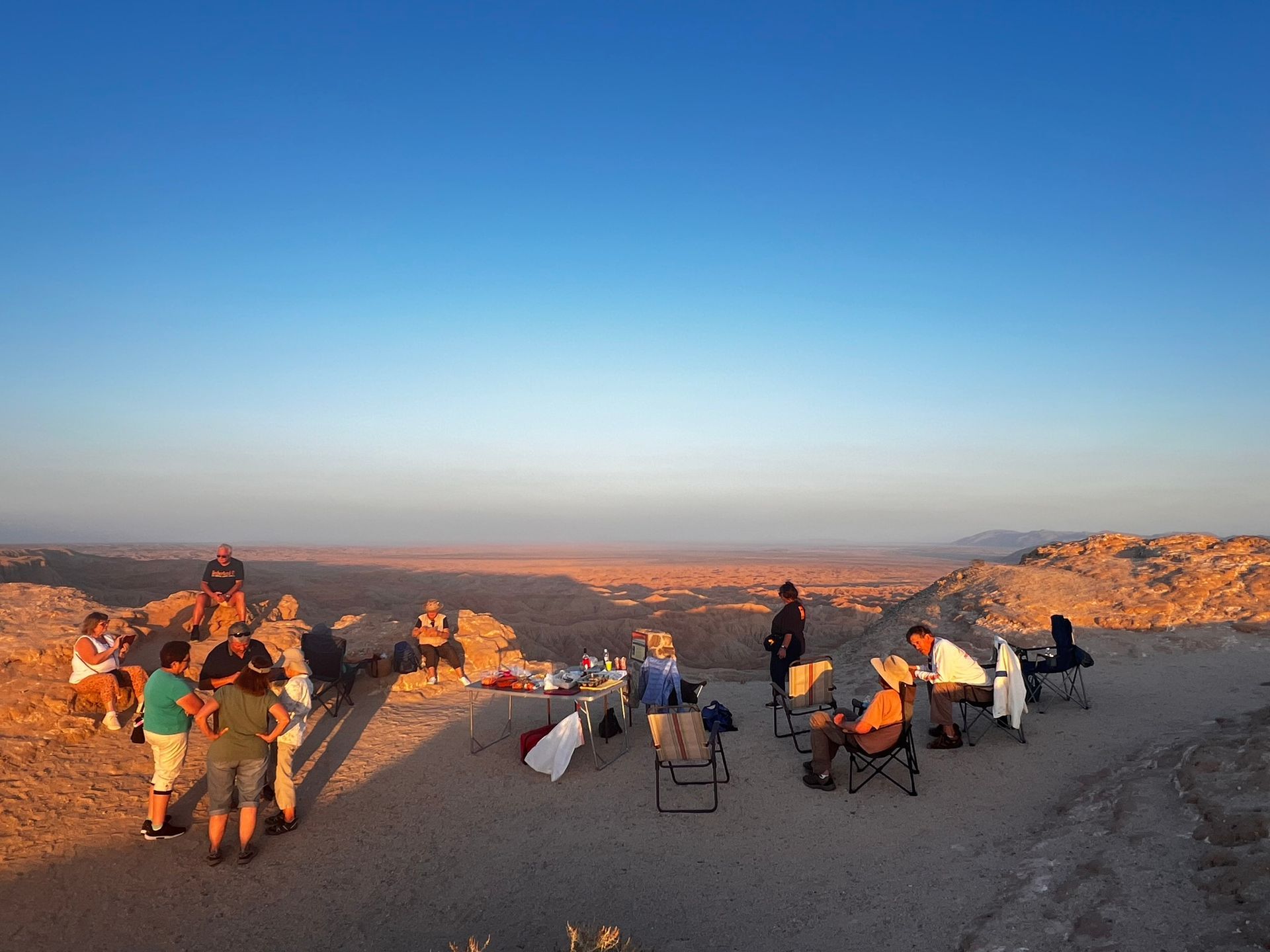People gathered on a rocky desert overlook at sunset, with chairs and bags under a clear blue sky.