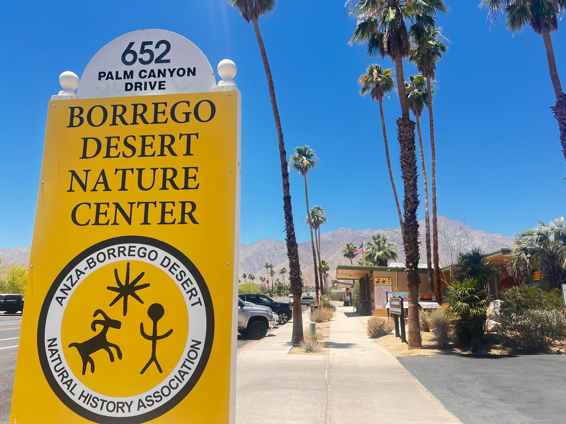 Borrego Desert Nature Center sign beside a sunny desert walkway with palm trees and mountains