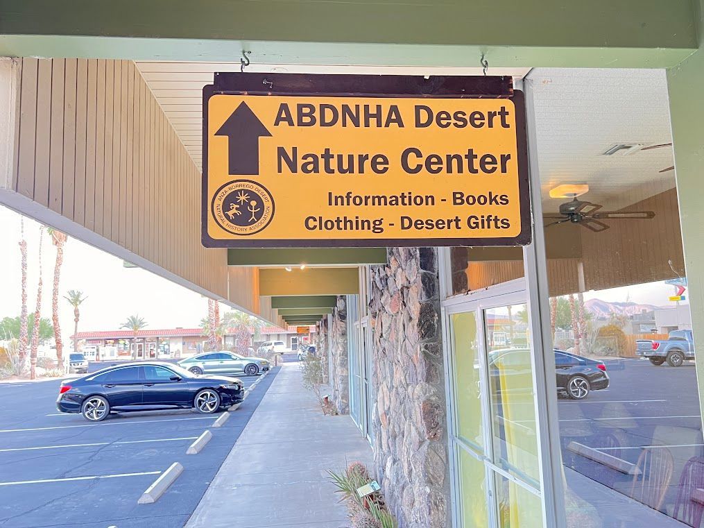 Yellow sign for ABDNHA Desert Nature Center with information, books, clothing, and desert gifts on a sidewalk.