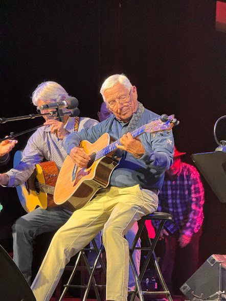 Two musicians seated on stage, playing acoustic guitars under dark lighting