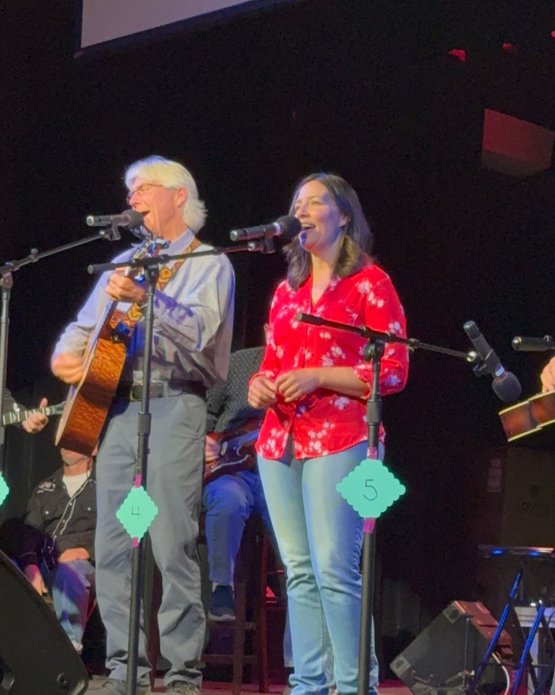 Two musicians singing on stage, one playing acoustic guitar, under dim concert lighting