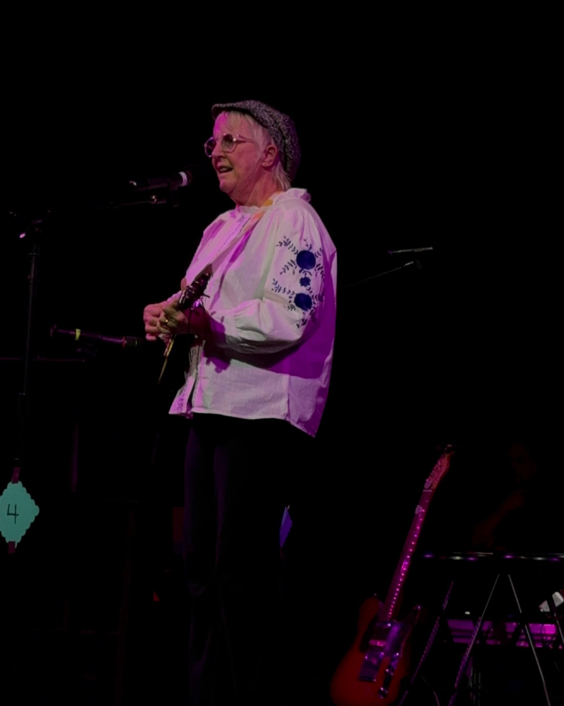 Performer in a white shirt playing guitar on a dark stage under pink-purple lights