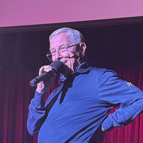 Man holding a microphone on stage under purple lighting, speaking or singing with one hand on his hip