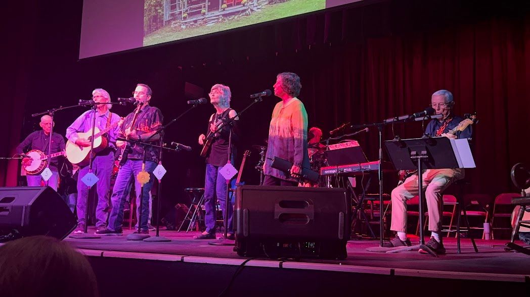 Six-piece band performing on a lit stage with purple and pink lighting, seated and standing with guitars and keyboards