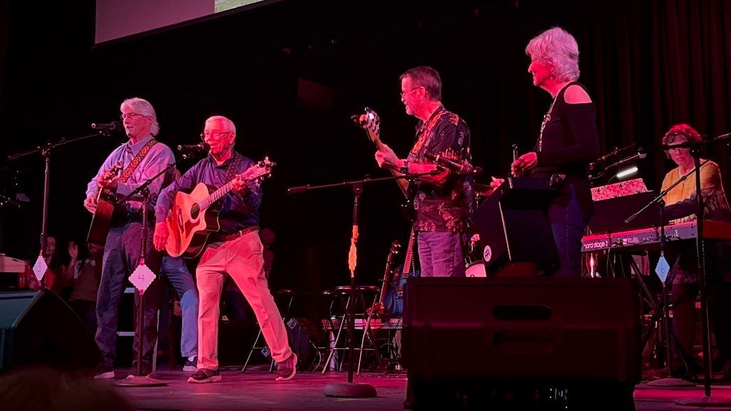 Band performing on a dimly lit stage with pink lighting, guitars, drums, and microphones