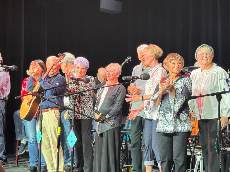 A choir of elderly singers stands on stage behind microphones, holding songbooks and facing the audience.