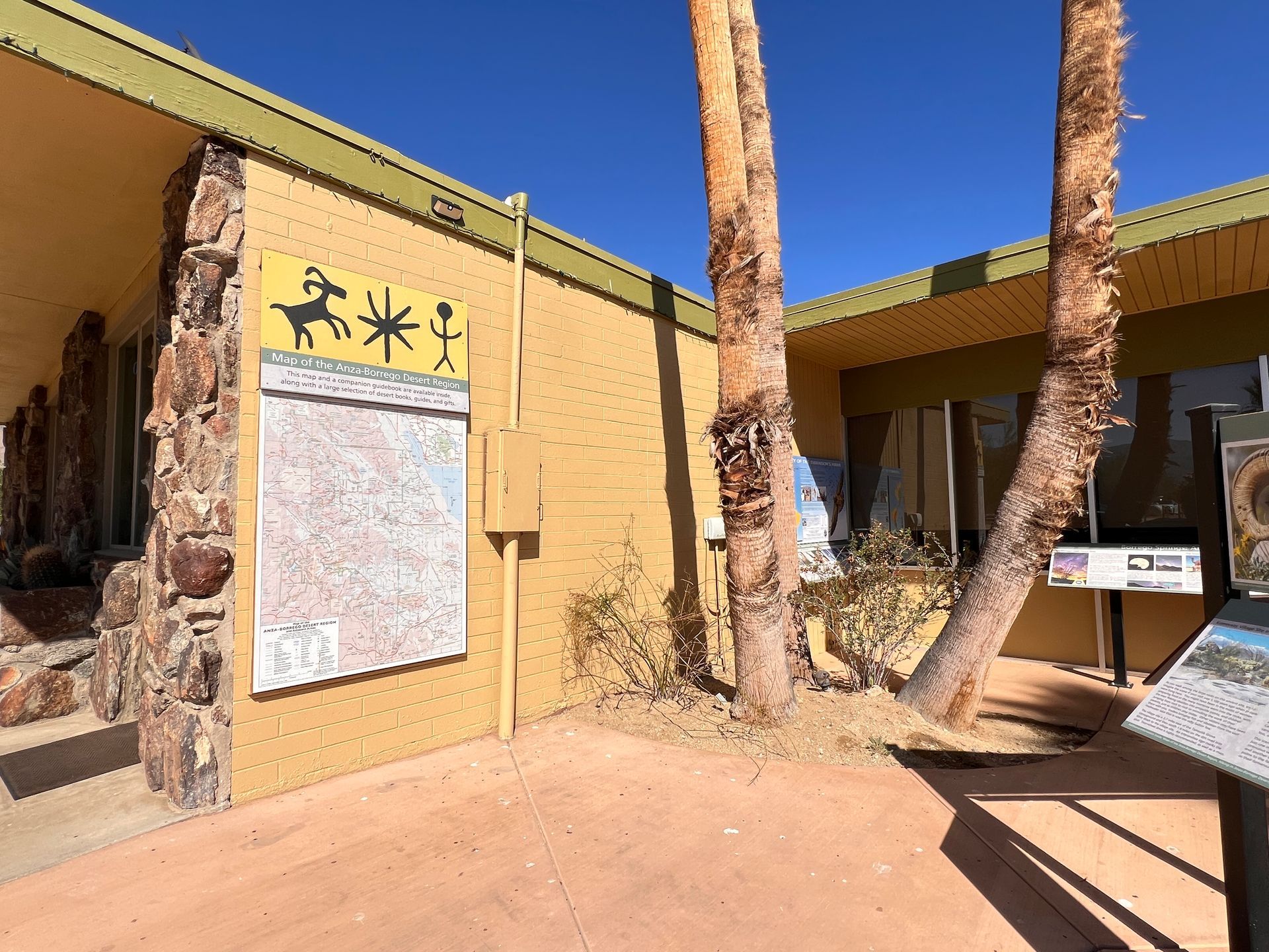 Desert museum entrance with tan walls, palm trees, and an informational signboard in bright sunlight