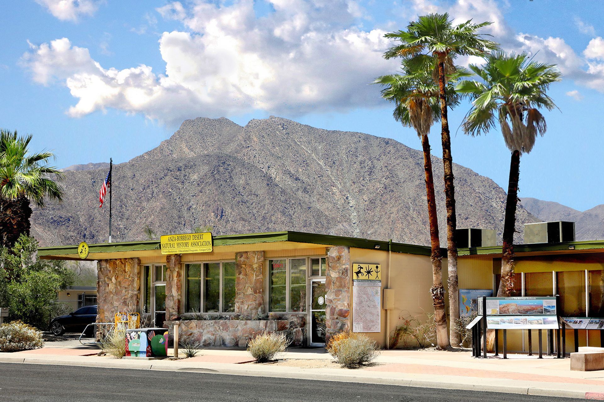 Small desert roadside building with palm trees and mountains under a cloudy sky