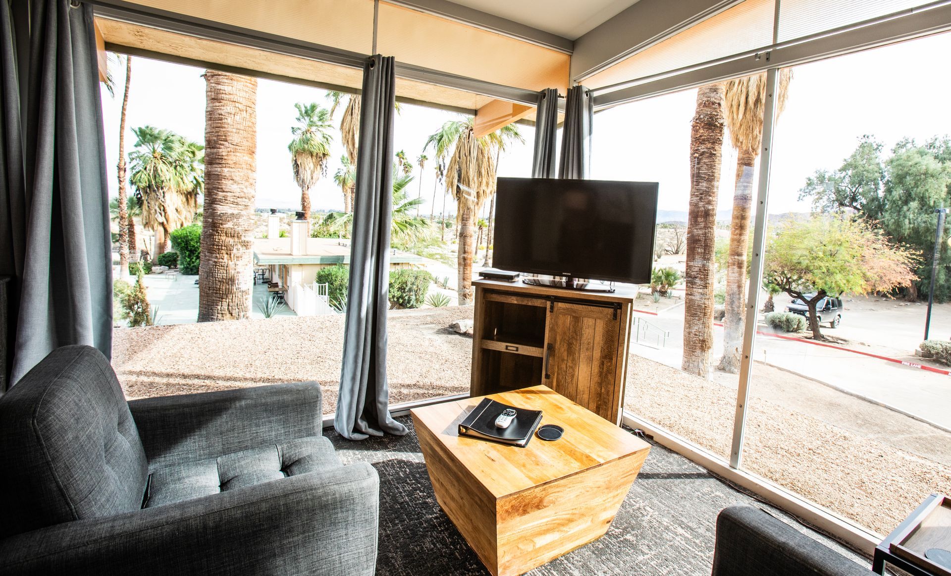 Bright living room with a gray couch, wooden coffee table, TV, and large windows overlooking palm trees.