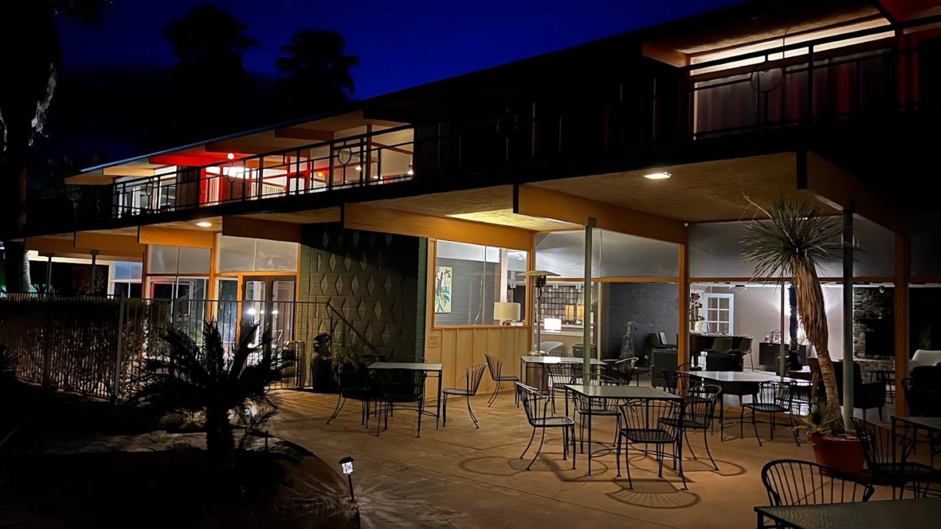 Outdoor restaurant patio at night with lit tables, chairs, and warm lights under a modern building overhang