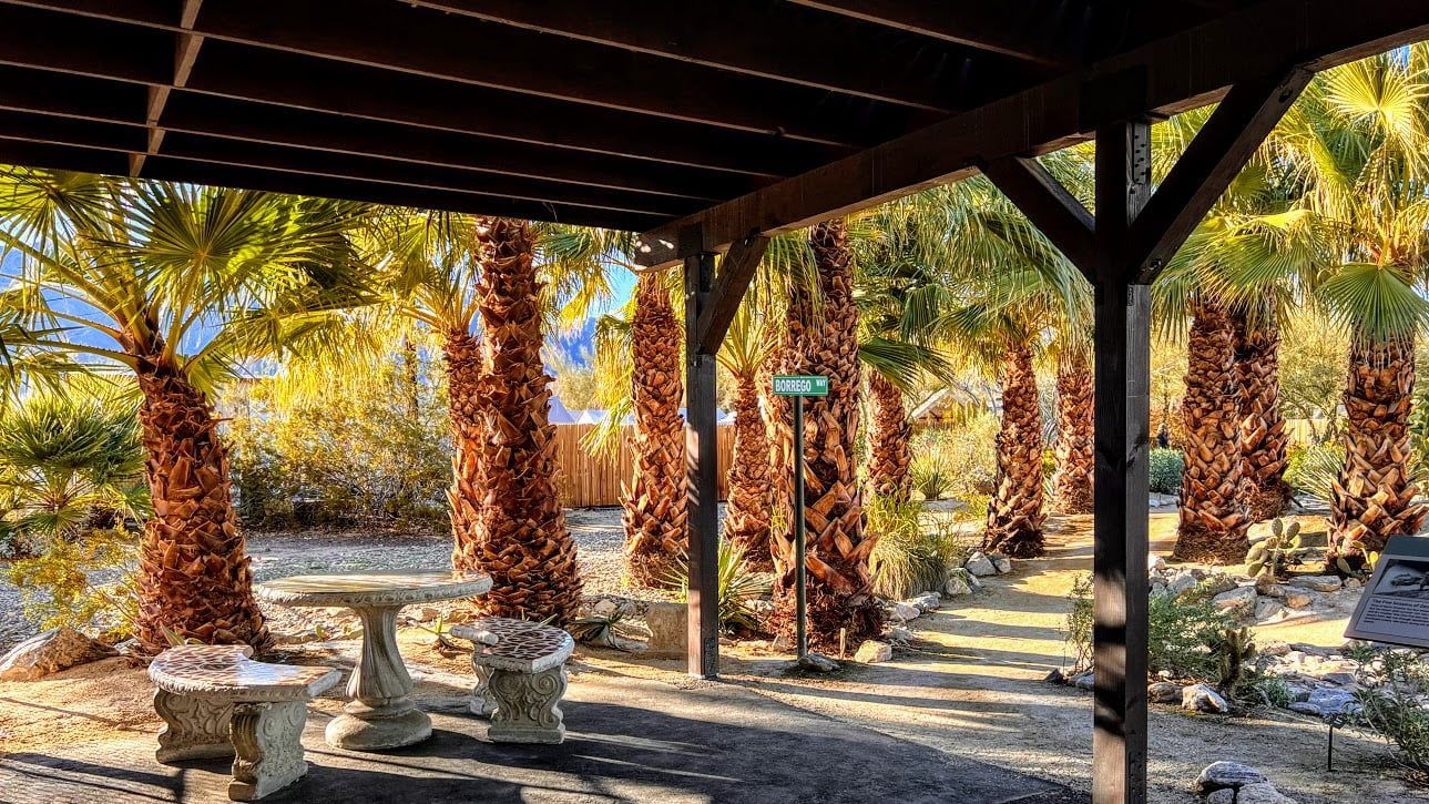 Shaded park shelter with stone benches overlooking rows of palm trees in a sunlit outdoor area