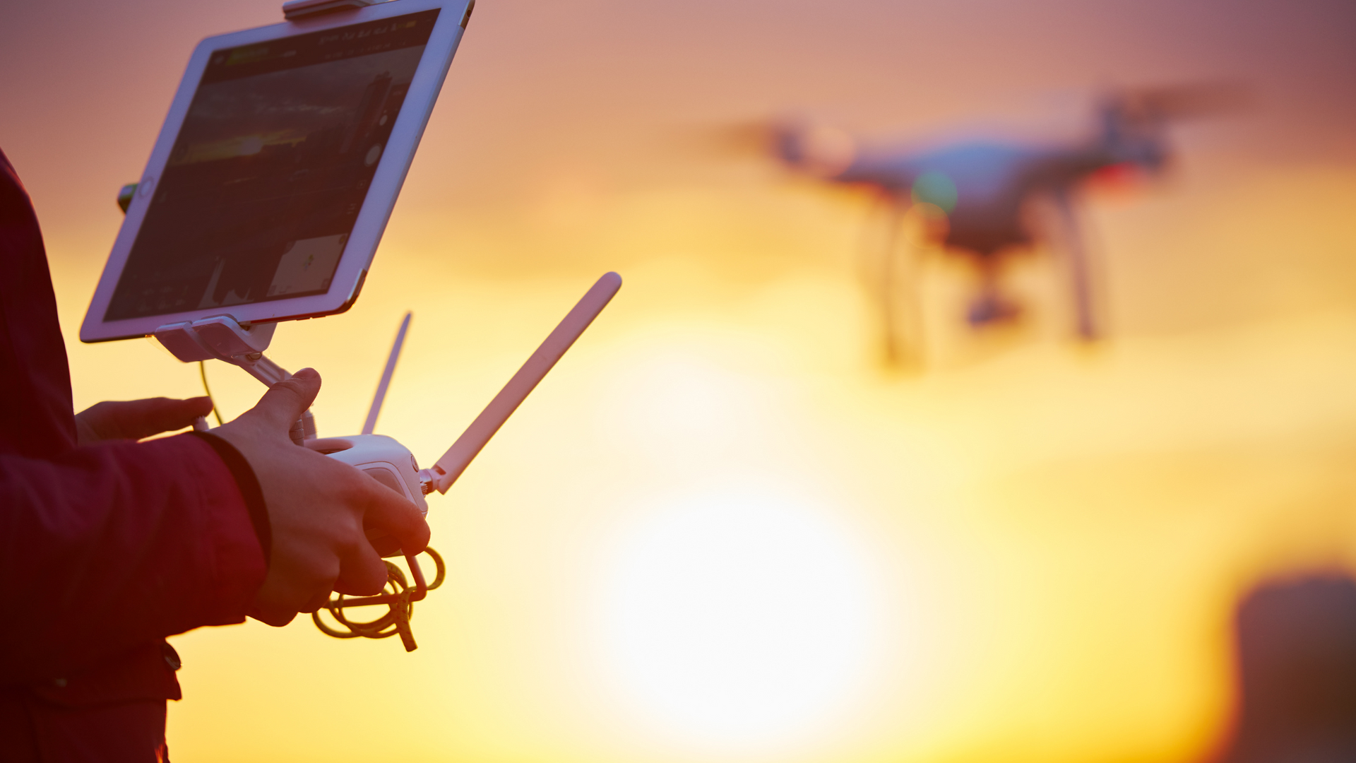 A person is holding a tablet and a remote control while a drone is flying in the background.