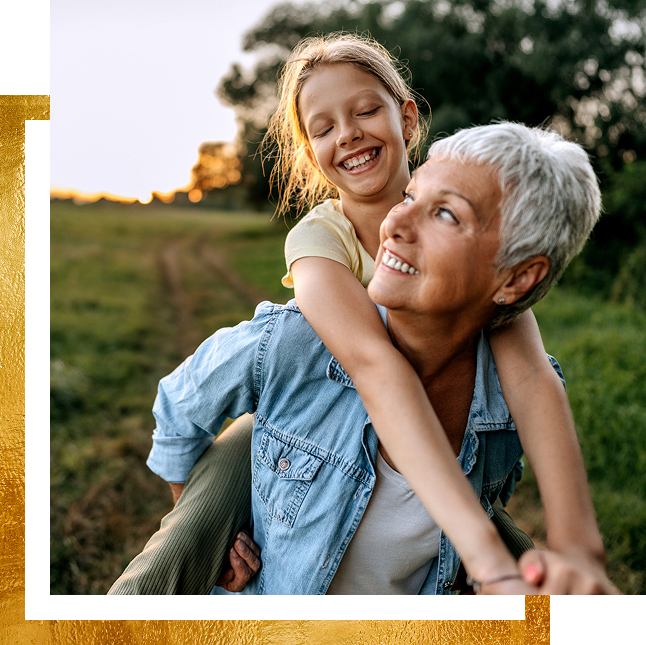 Grandmother gives piggyback ride to granddaughter outdoors, both smiling.