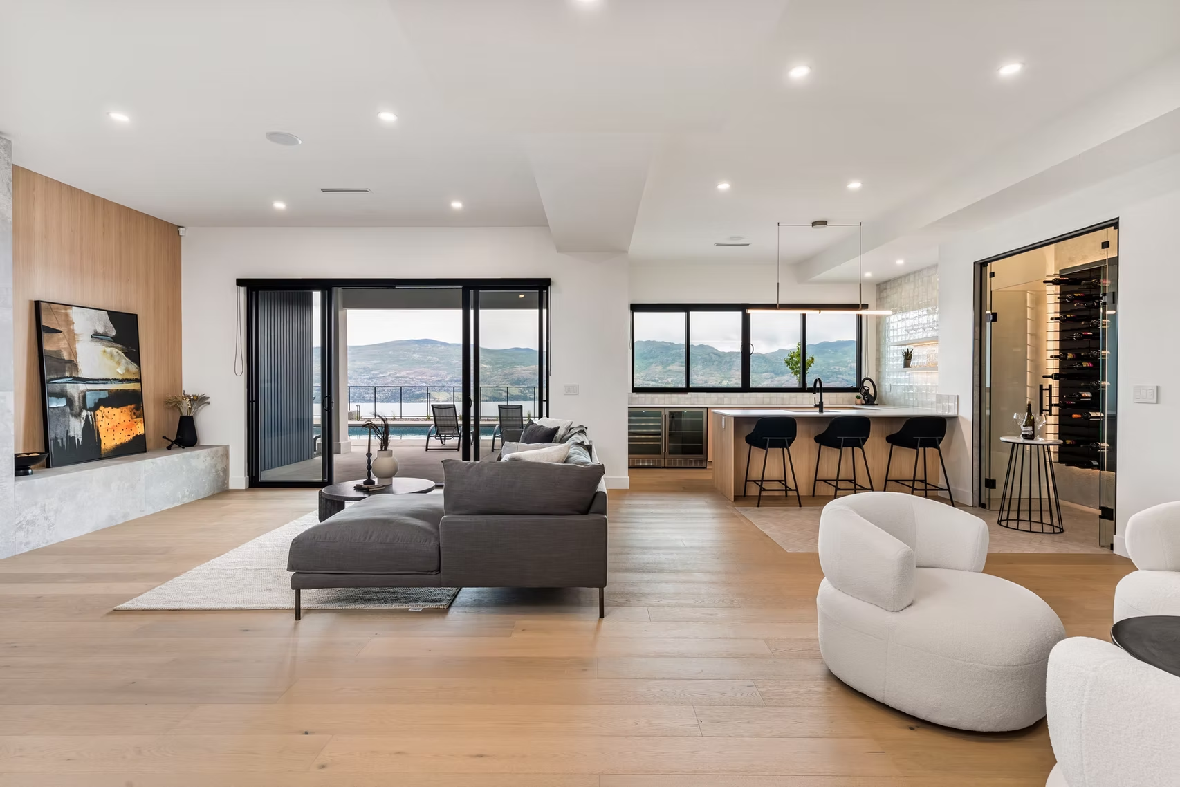 Modern open-plan living room with gray sofa, white chairs, wood floors, and large sliding glass doors to a view.