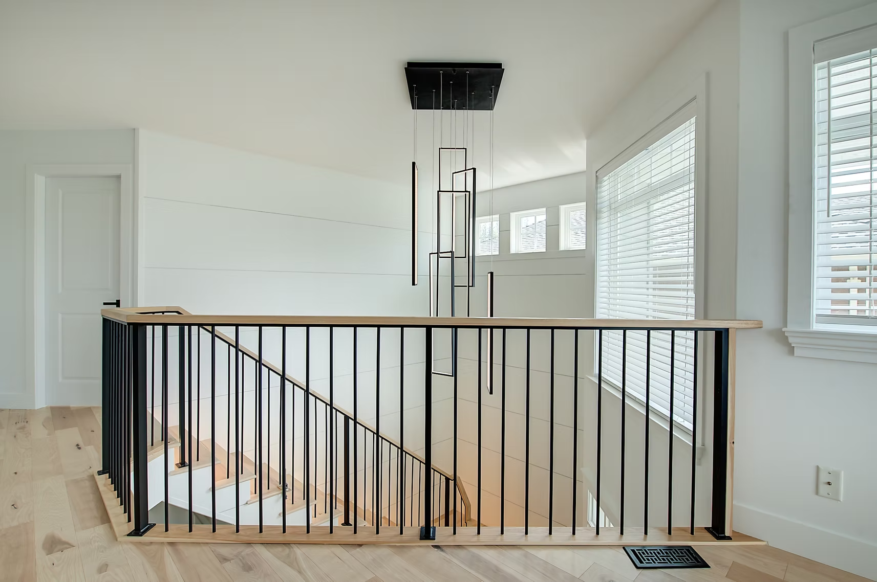 Modern hallway with black stair railing, light wood floors, and white walls and windows