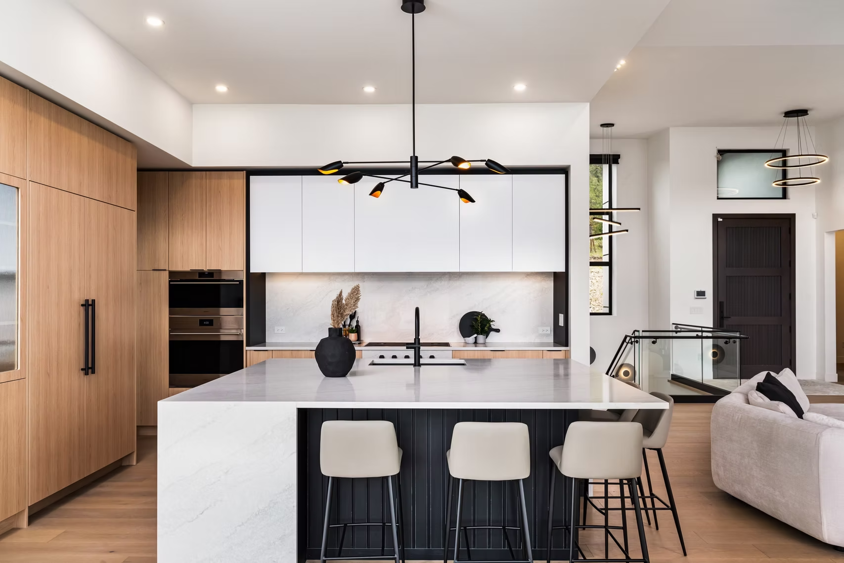 Modern kitchen with wood cabinetry, white island, black pendants, and light gray stools in an open-plan home