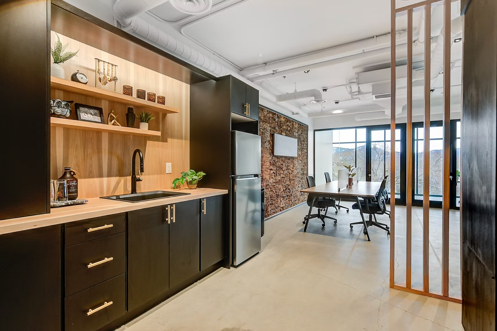 Modern office kitchen with wood cabinetry, black appliances, and a glass-walled meeting area beyond