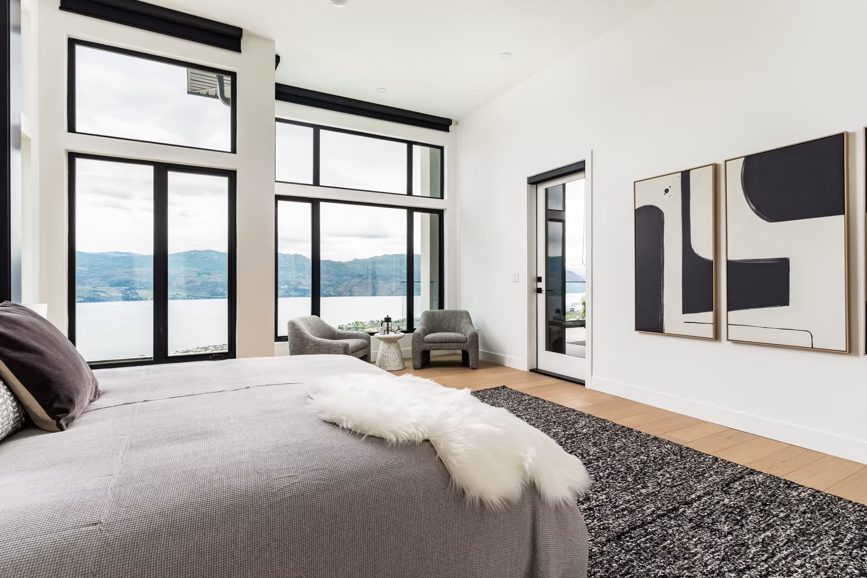 Modern bedroom with large windows, gray bedding, abstract wall art, and a lake view
