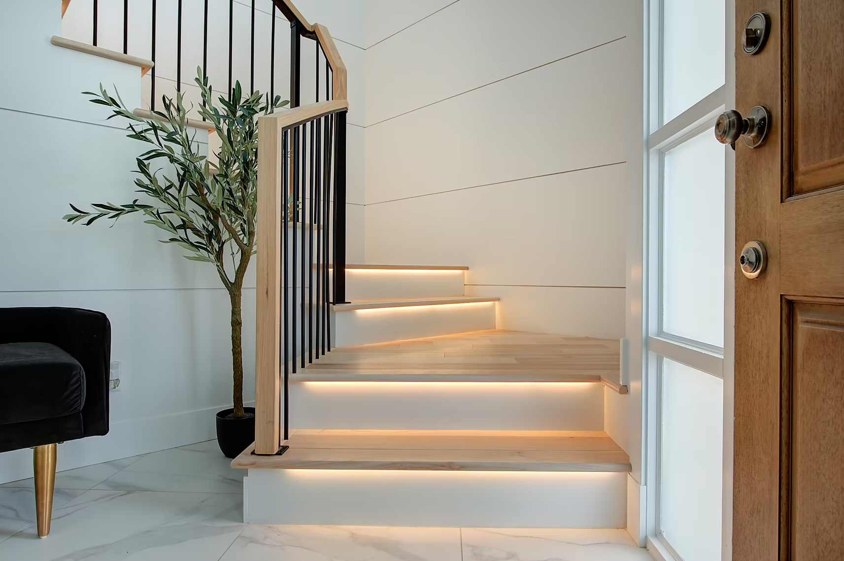 Modern staircase with glowing step lights, black railing, potted plant, and a wooden front door