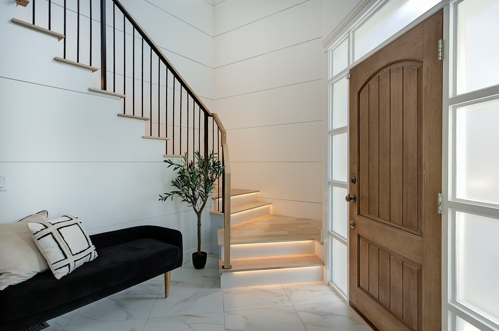 Bright foyer with white stairs, wooden front door, black bench, and potted plant