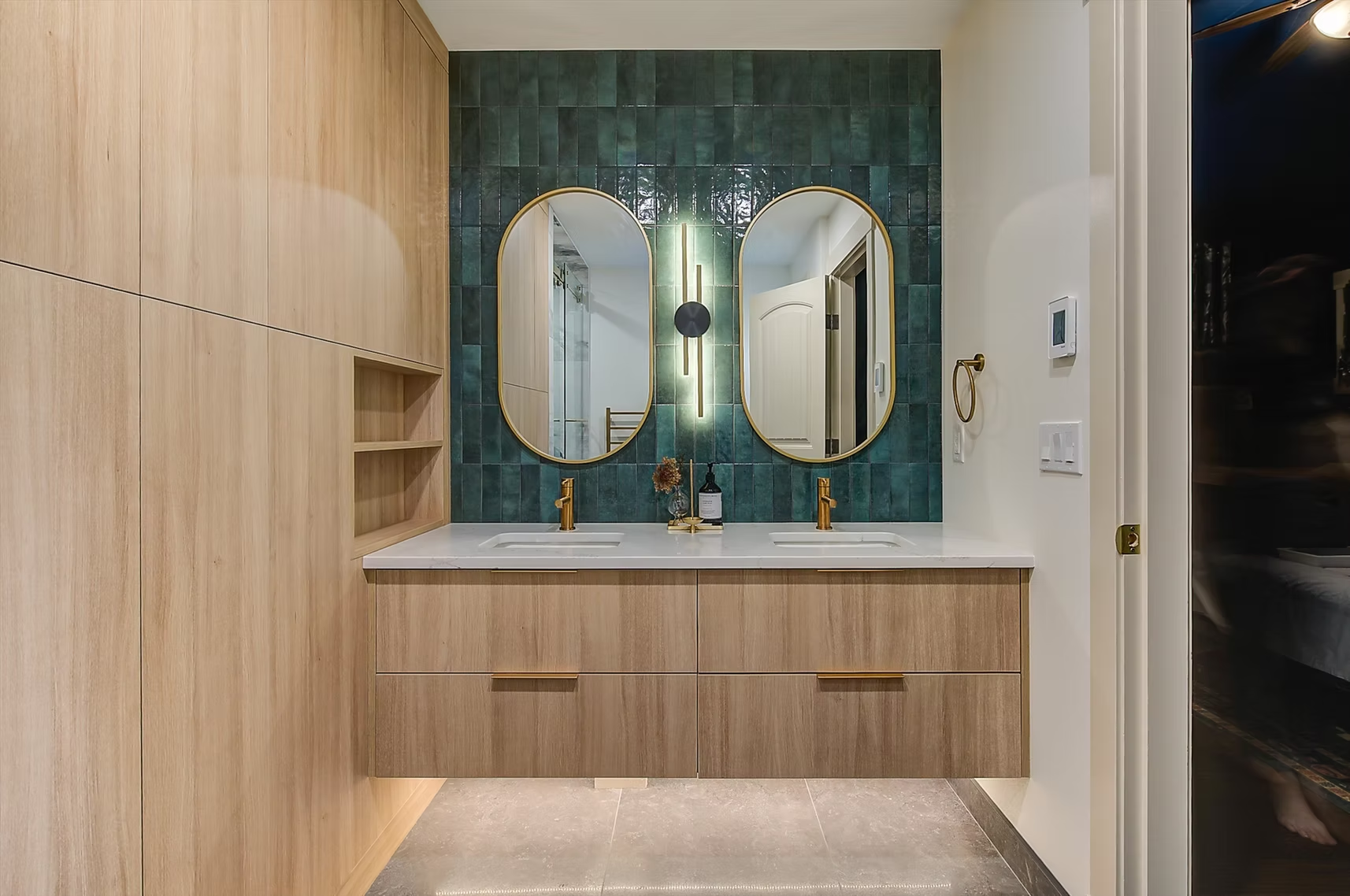 Modern bathroom vanity with twin oval mirrors, wood cabinetry, marble counter, and green tiled accent wall.
