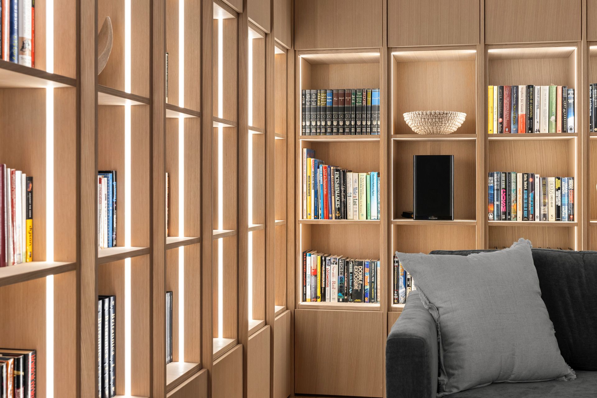 Warm wood-paneled library with built-in bookshelves and a gray sofa by a large window.