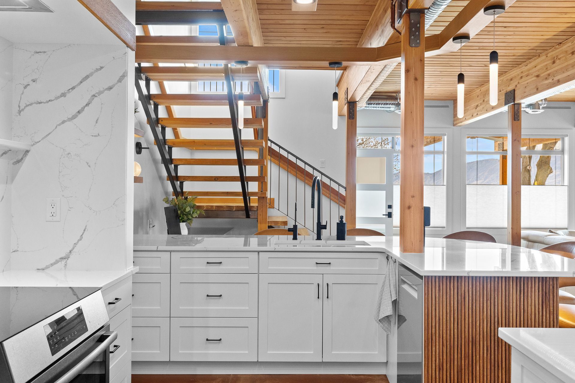 Modern kitchen with white cabinets, marble backsplash, wood beams, and stairs leading to a loft.