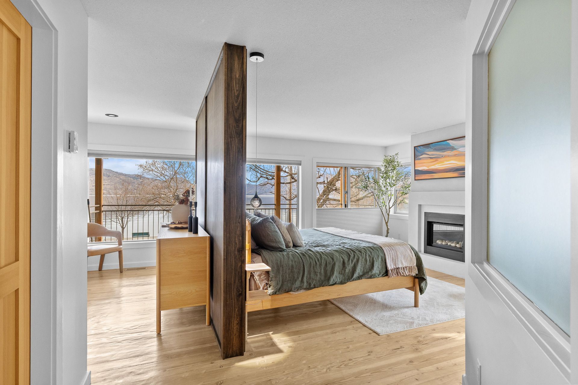 Bright modern bedroom with a wooden bed, frosted glass wall, and large windows overlooking trees