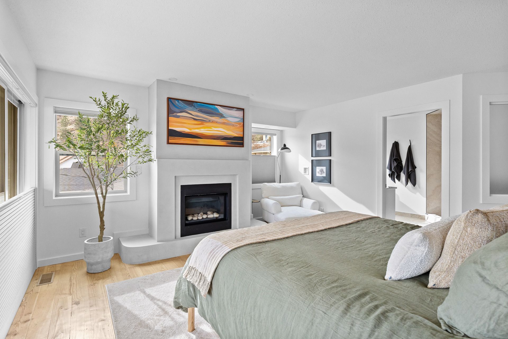 Bright gray bedroom with a bed, fireplace, wall art, and large windows with a potted tree