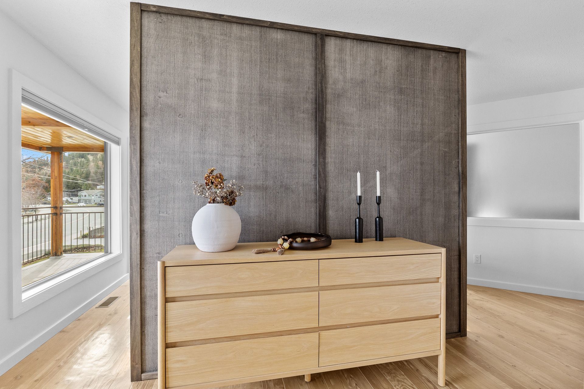 Modern bedroom with wooden dresser, gray panel wall, and bright windows