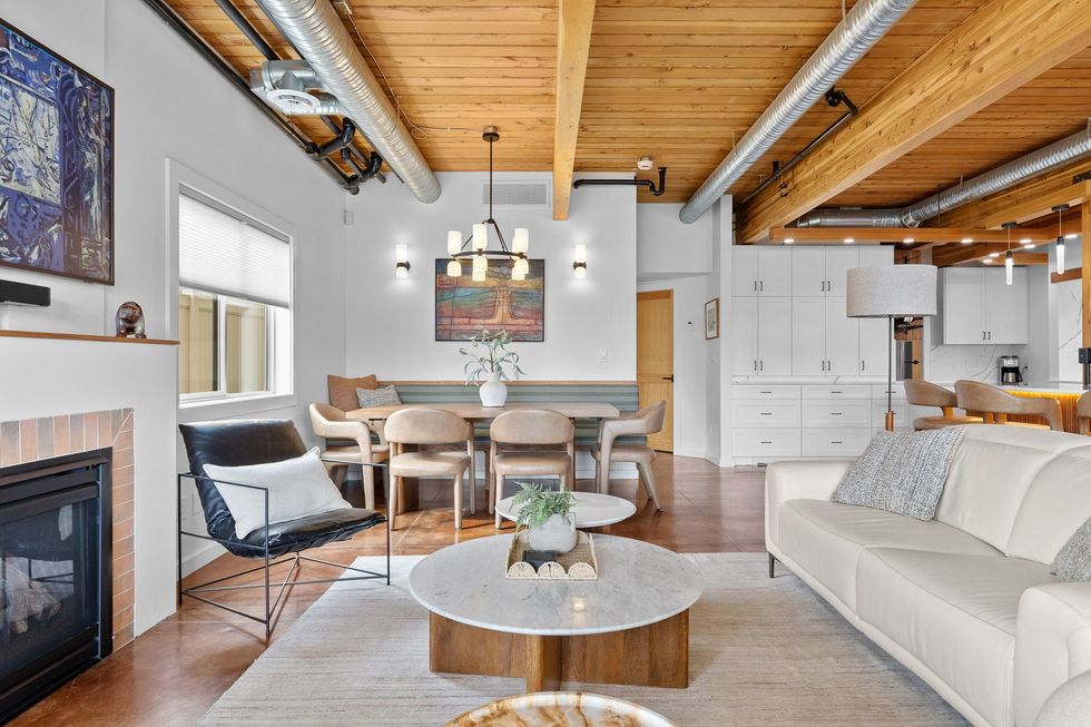 Bright open-concept living room with wood ceiling beams, fireplace, white sofa, dining table, and kitchen area