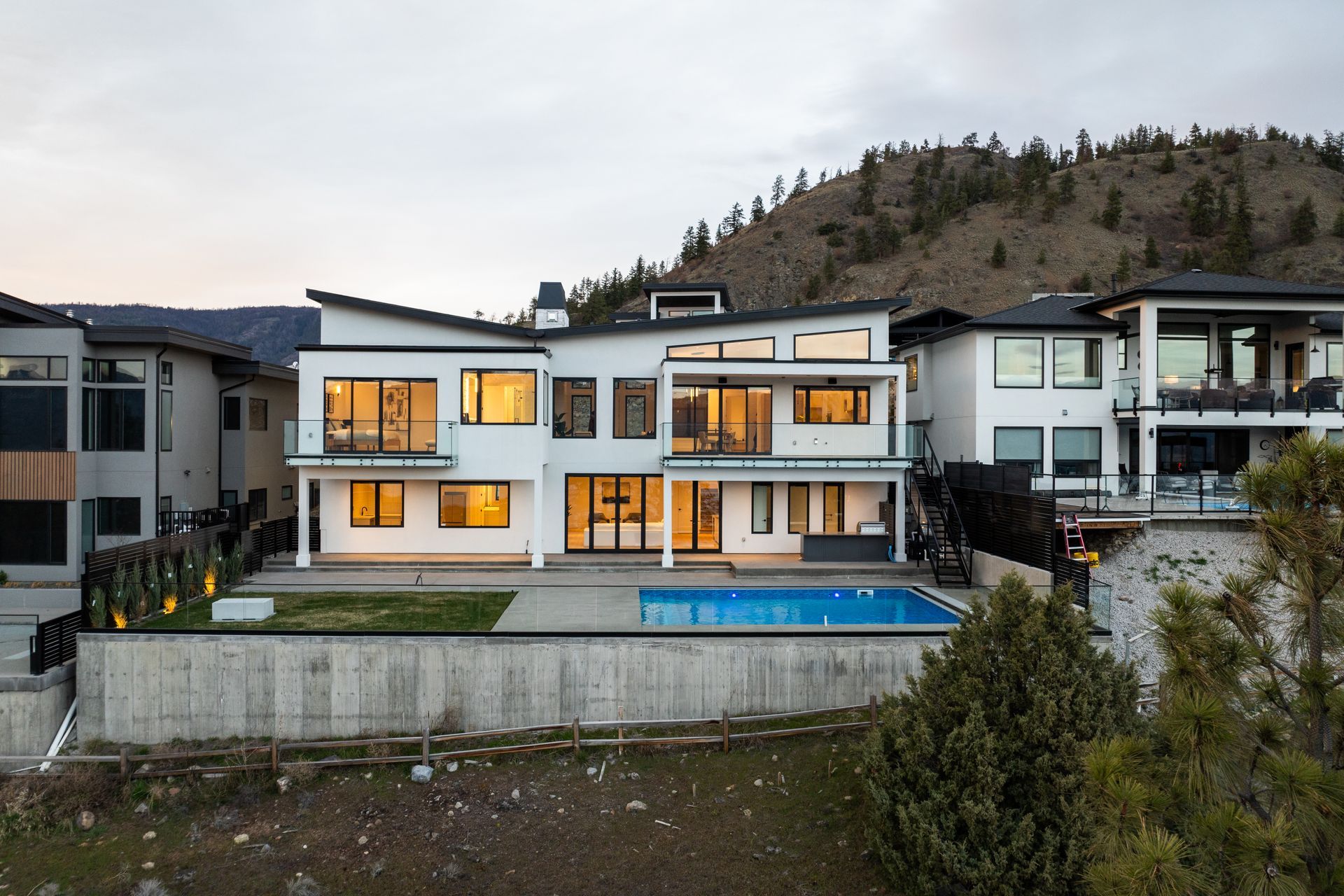 Modern waterfront house with lit windows, a pool, and hillside backdrop at dusk