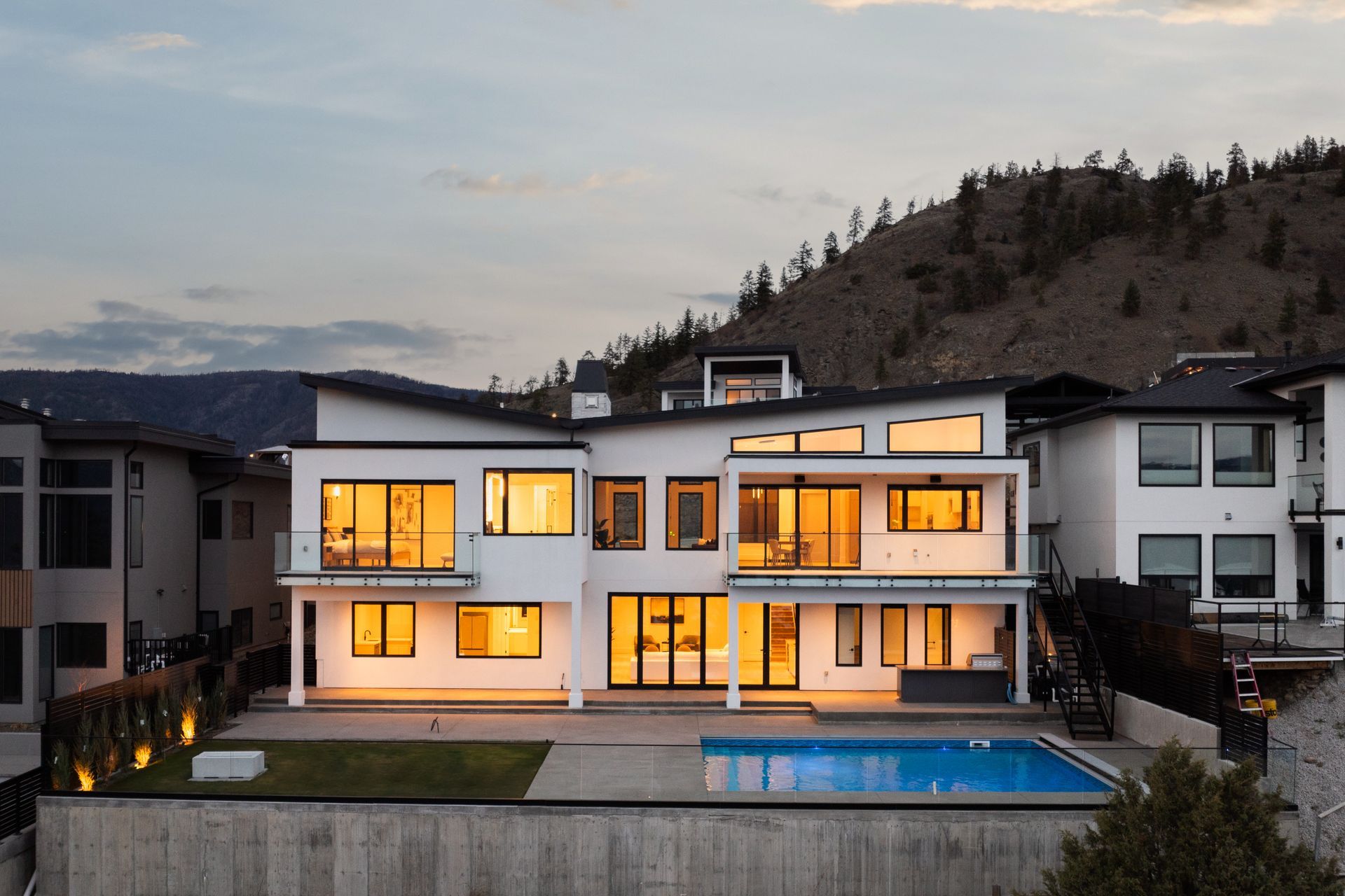 Modern white hillside home at dusk with warm lights and a backyard pool