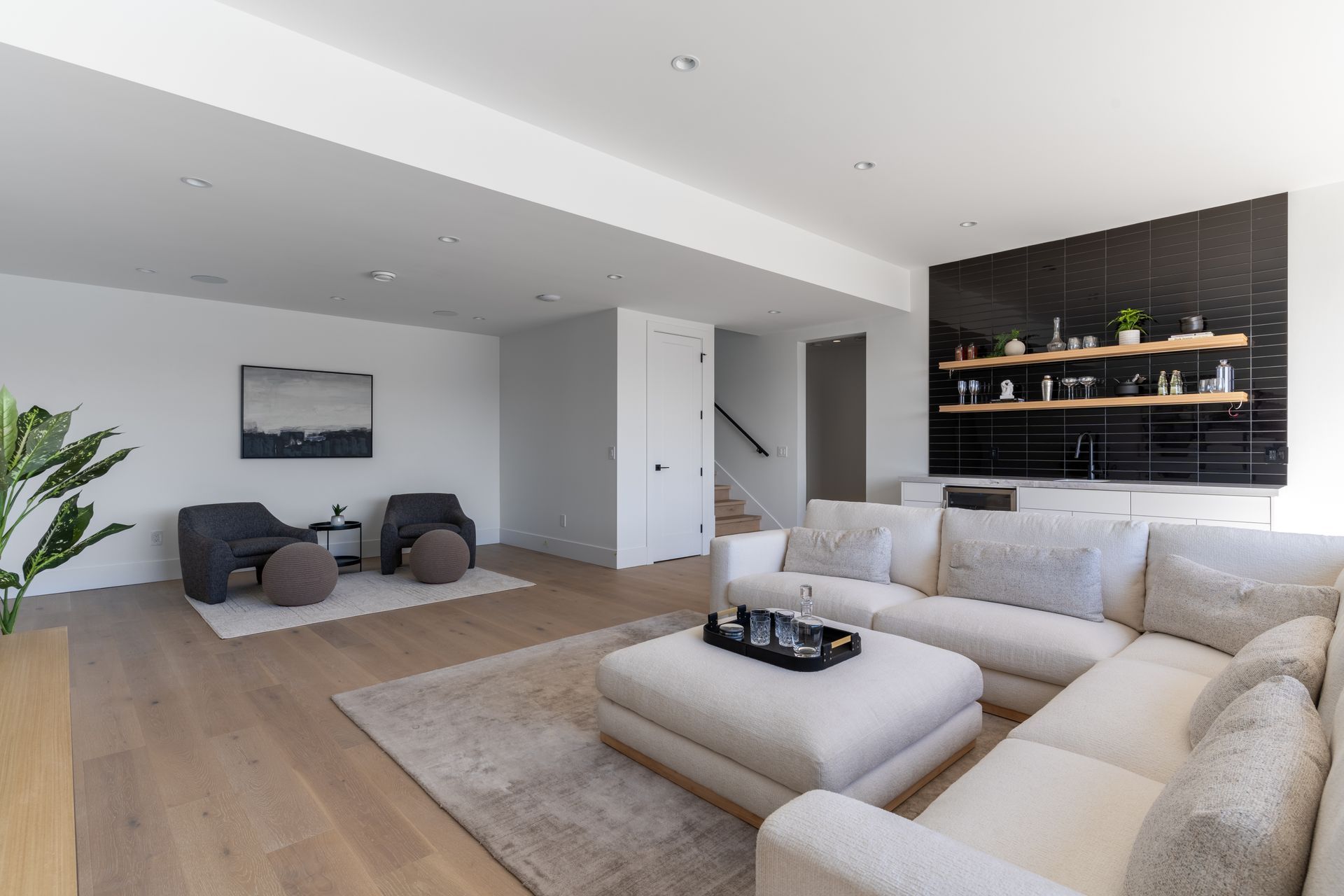 Bright modern living room with beige sectional, dark accent wall, and lounge chairs by a large window
