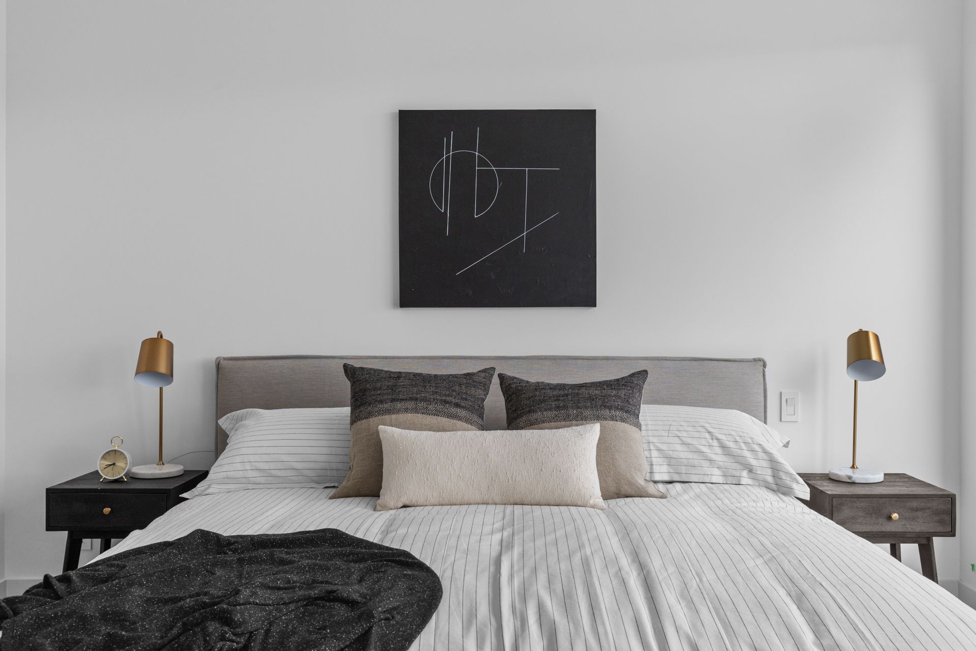 Minimalist bedroom with gray bed, white bedding, black wall art, and gold bedside lamps.
