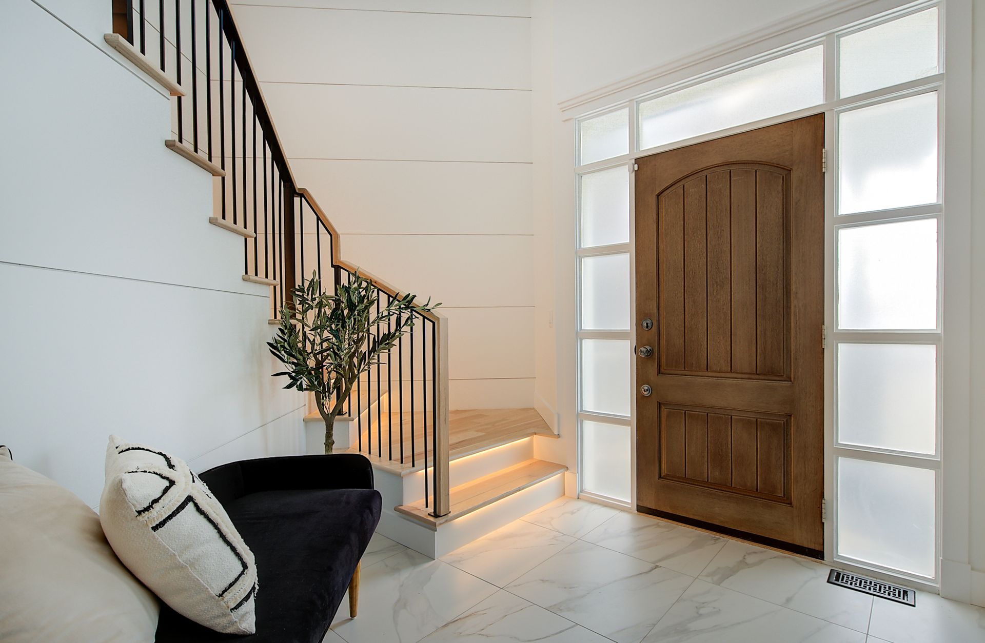 Bright entryway with wooden front door, white walls, staircase, and bench with pillows and plant.