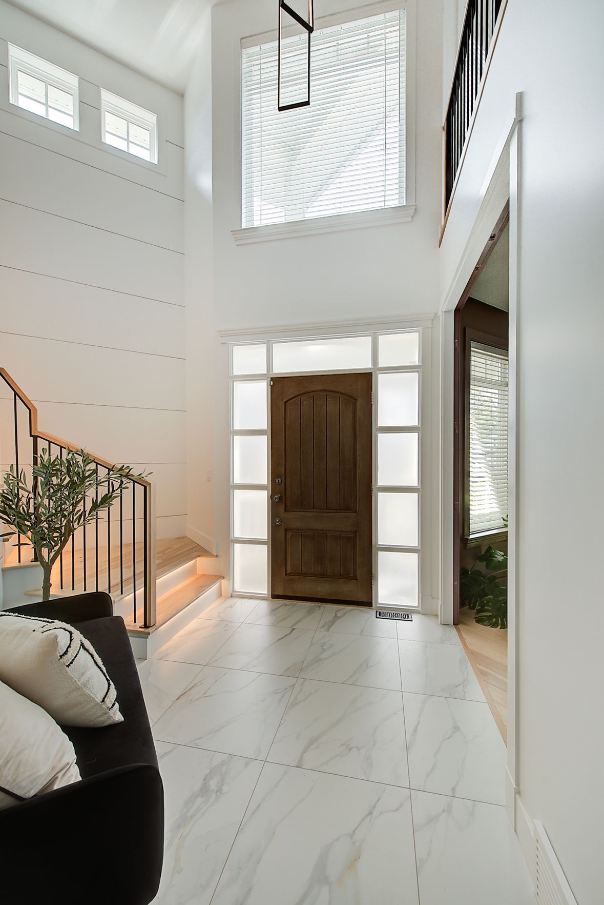 Bright, modern foyer with white walls, marble floor, wooden front door, and staircase with warm lighting