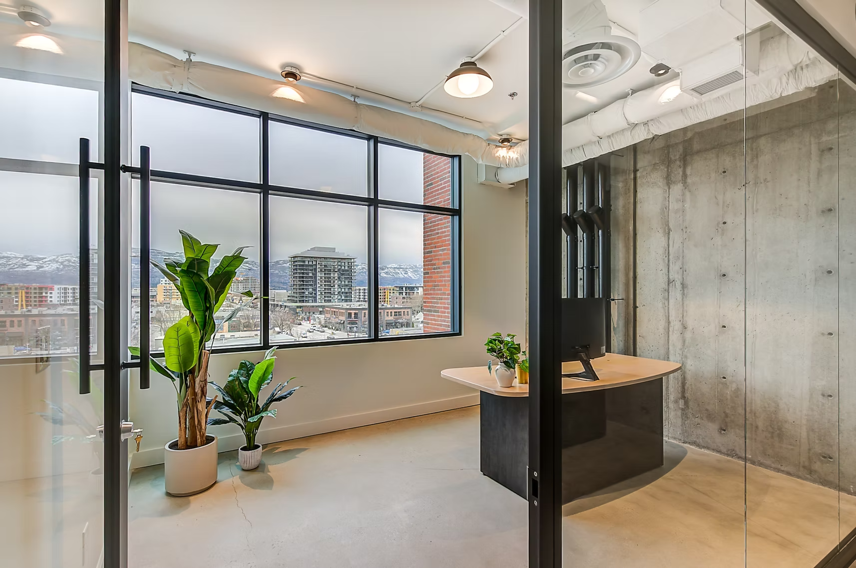 Modern office with large windows, concrete wall, wooden desk, and potted plants overlooking a city view