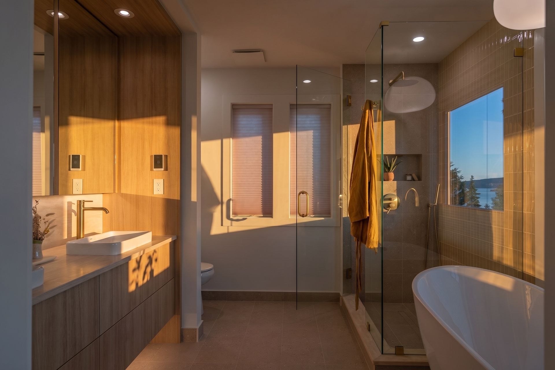 Modern bathroom with wood cabinetry, glass shower, bathtub, and warm sunset light.