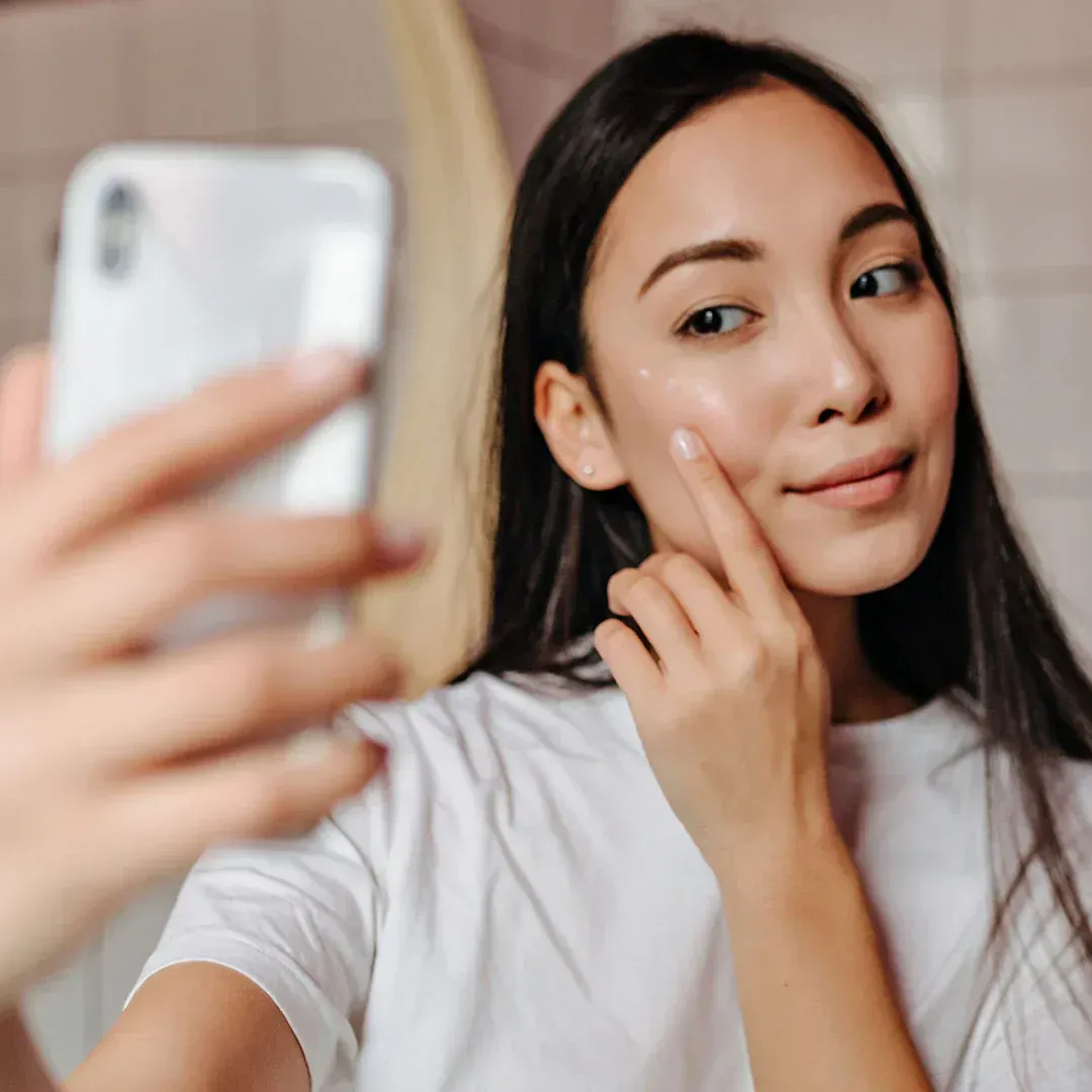 Woman focusing on skin on face in a mirror , taking a selfie, touching her cheek in a bright room.