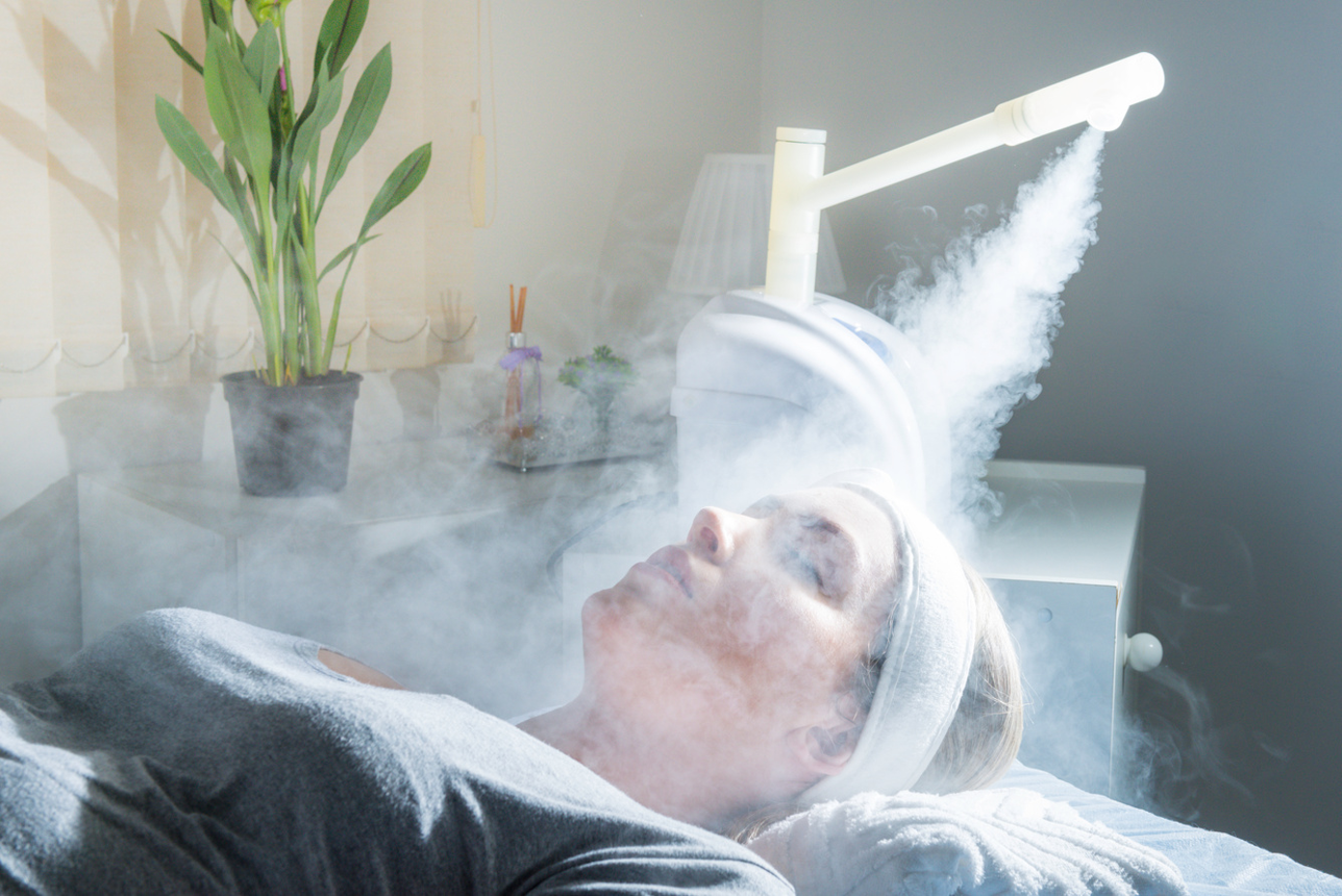 Person reclining under a steaming facial spa device in a bright room