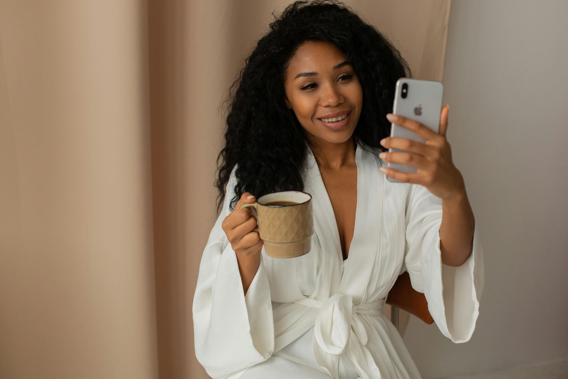 Person in a white robe smiling and taking a selfie while holding a mug indoors
