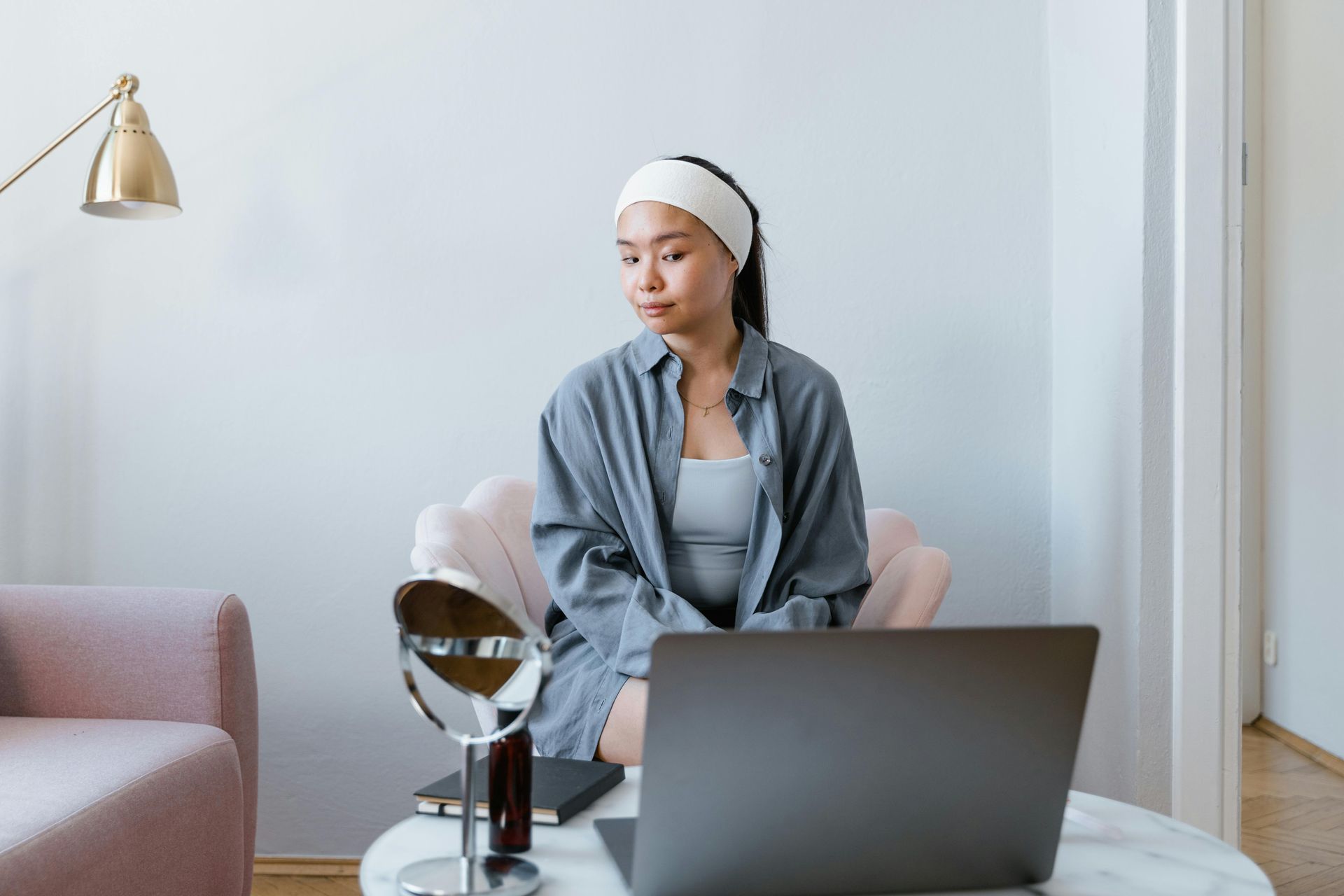 Woman wearing a facial headband on her head using a laptop at a table in a bright living room looking into table top mirror