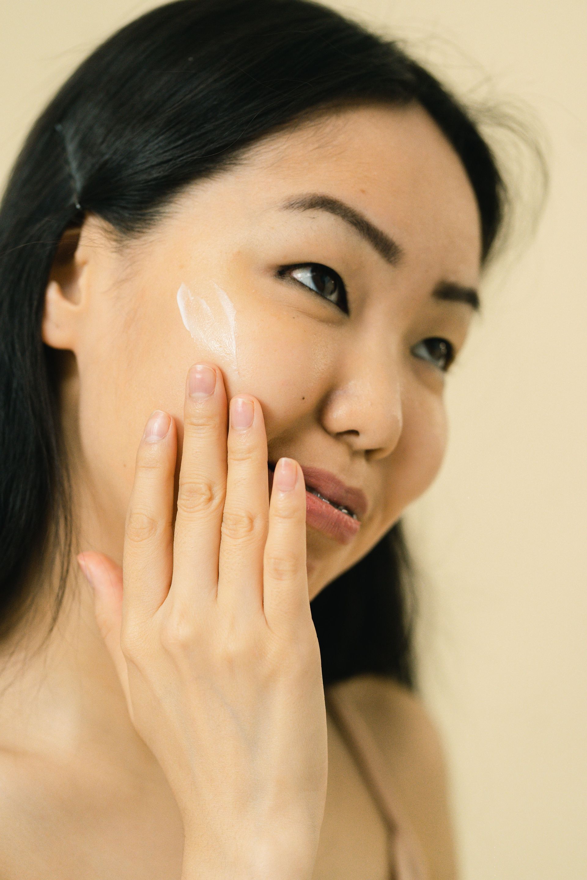 Woman applying cream to her cheek with a calm expression on a beige background