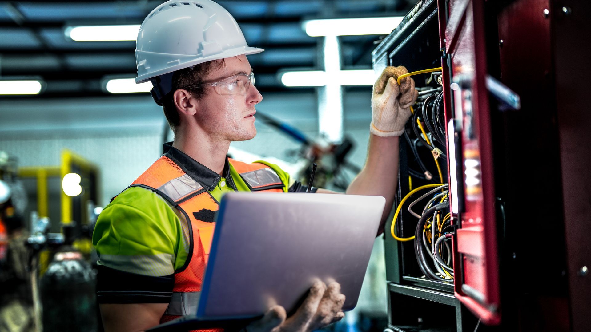 A male engineer in a hard hat inspects electrical wires while holding a laptop.