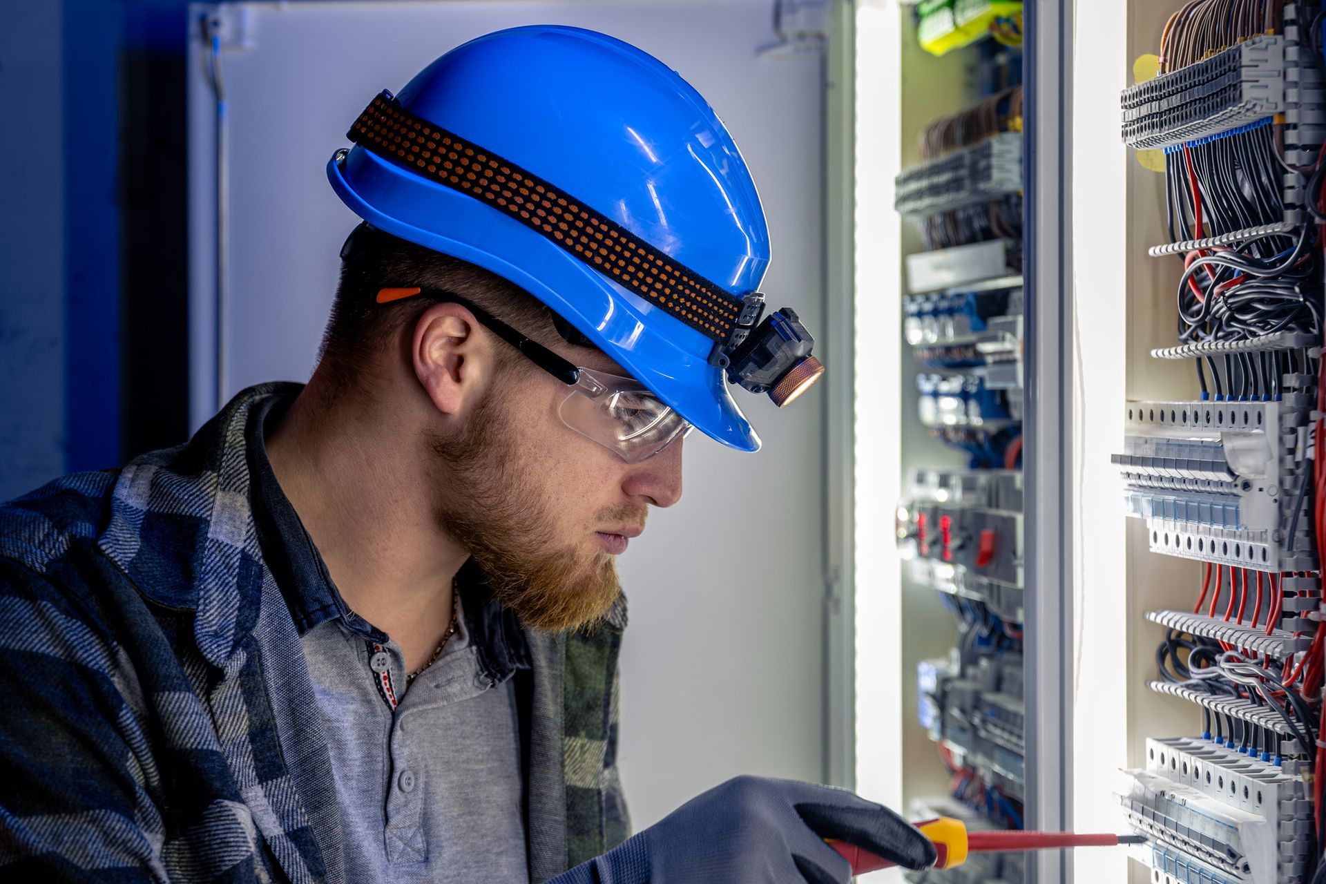 Electrician inspecting electrical control panel wiring with safety helmet and insulated screwdrivers.