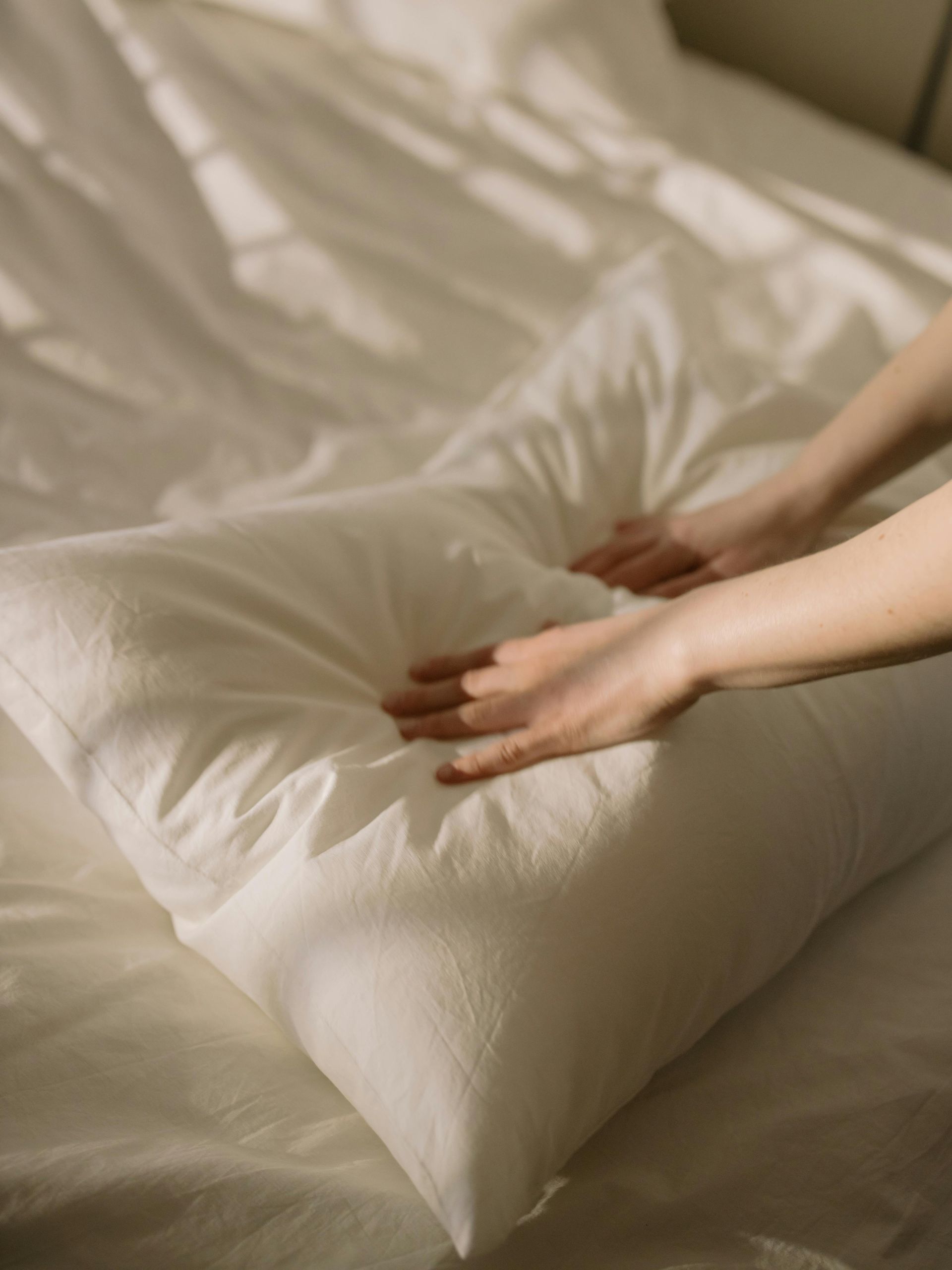 Hands smoothing a white pillow on a bed with white sheets, sunlight streaming in.