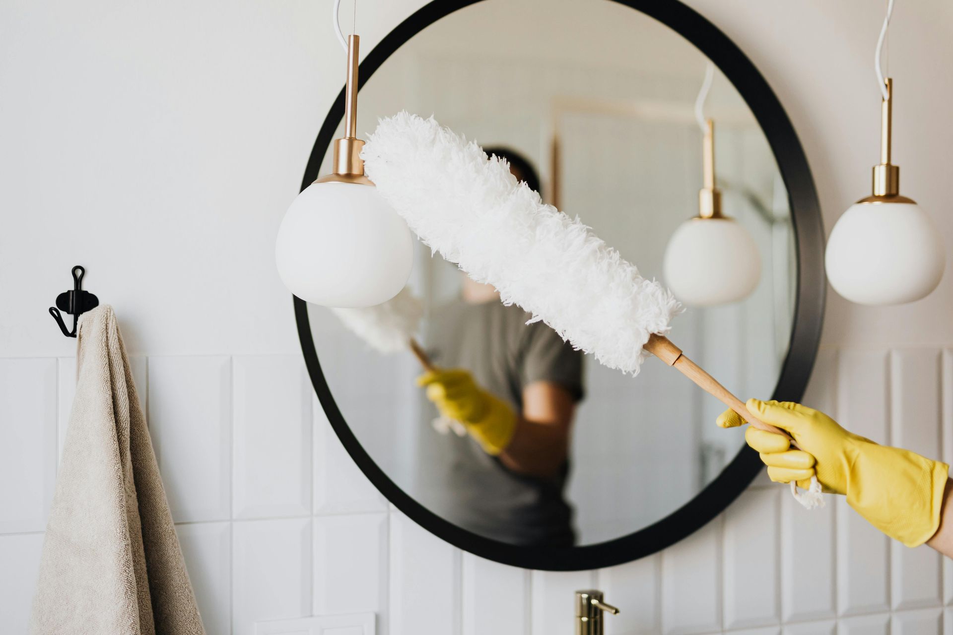 Person dusting a round mirror with a feather duster, wearing yellow gloves in a bathroom.