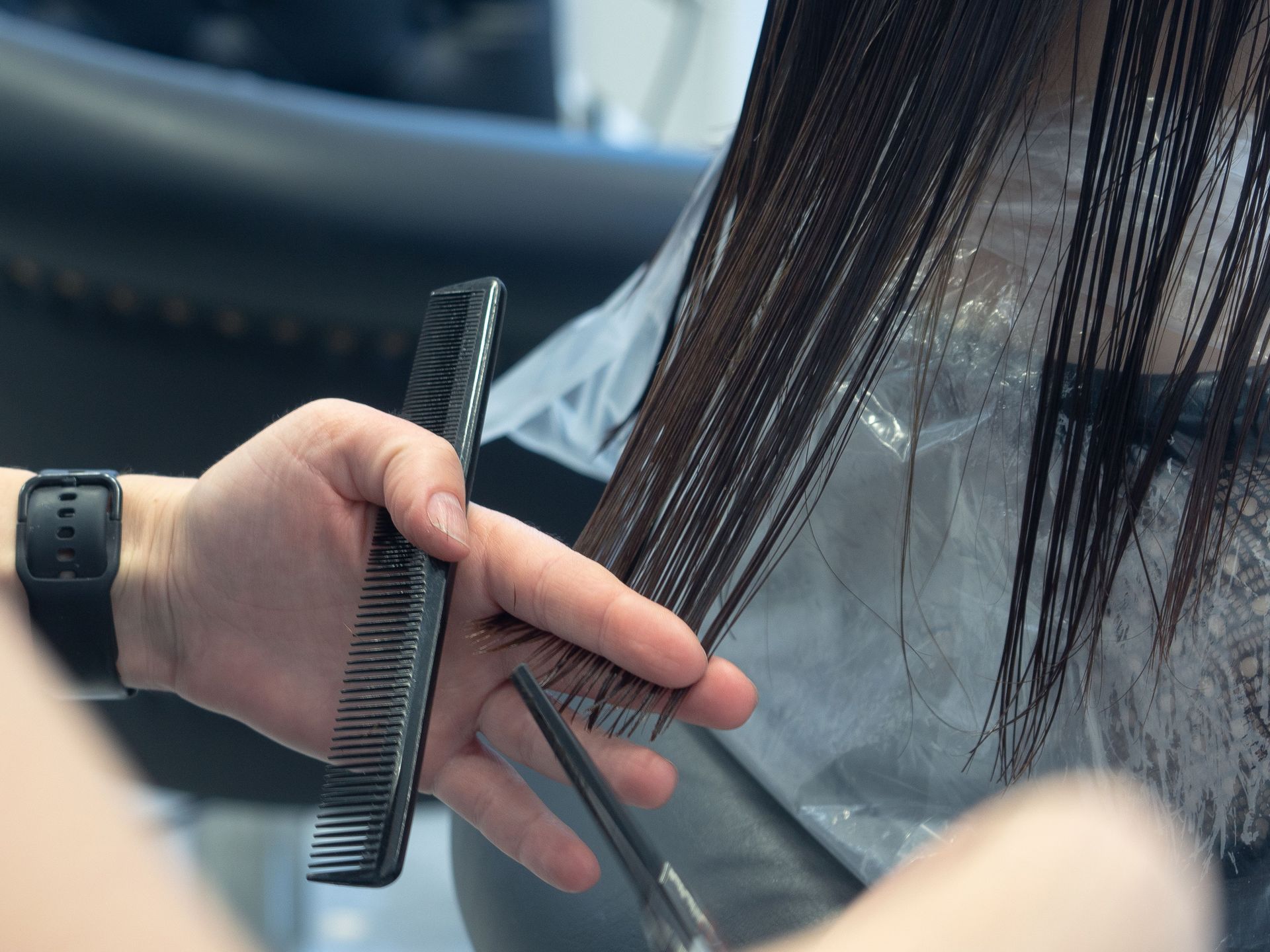 Hand holding a comb and cutting hair with scissors beside long dark strands in a salon