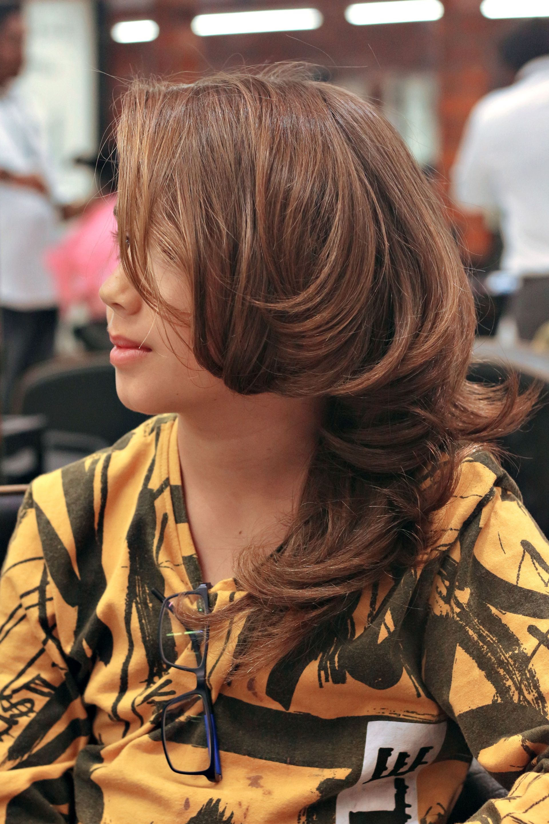 Woman with long brown hair in a yellow patterned top, seated in profile at an indoor event
